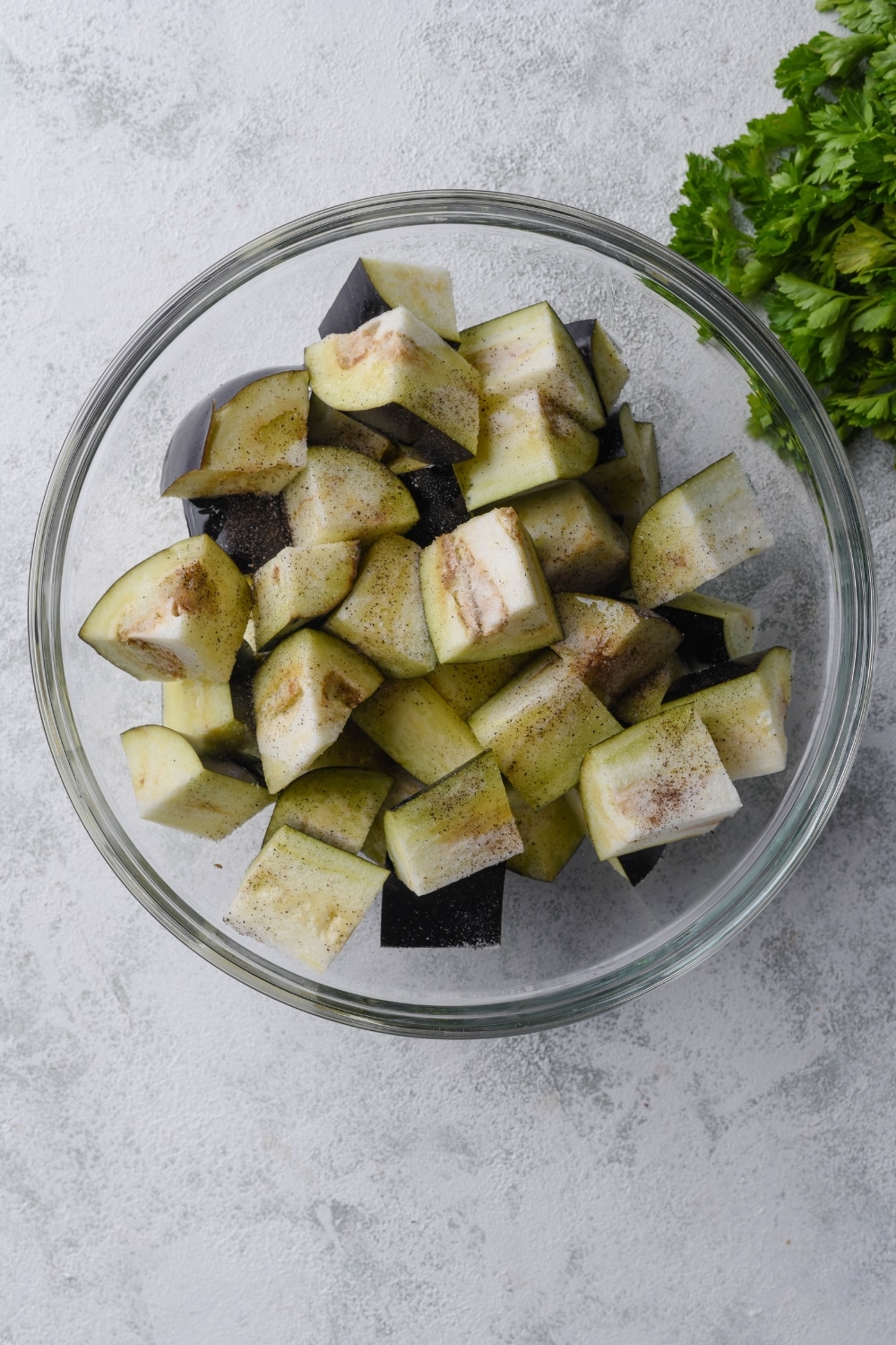 A glass bowl of uncooked eggplant chunks seasoned with salt and pepper.