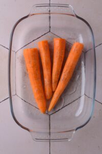 Four peeled and seasoned carrots on a glass baking dish.