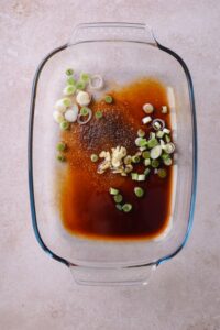 A glass baking dish with soy sayce, rice vinegar, brown sugar, sliced garlic cloves, and chopped green onions.