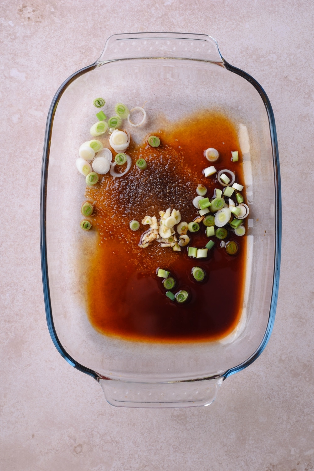 A glass baking dish with soy sayce, rice vinegar, brown sugar, sliced garlic cloves, and chopped green onions.