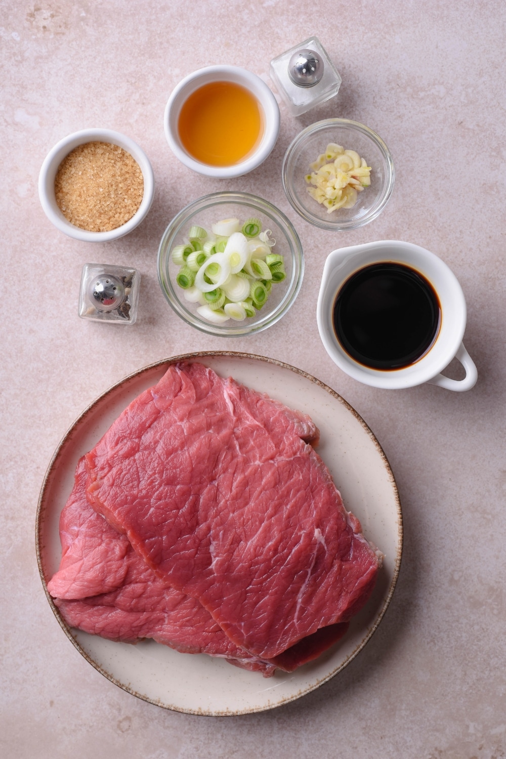 Raw sirloin tip steak on a plate, small bowls of soy sauce, rice vinegar, brown sugar, sliced garlic gloves, and sliced green onion, and salt and pepper shakers.