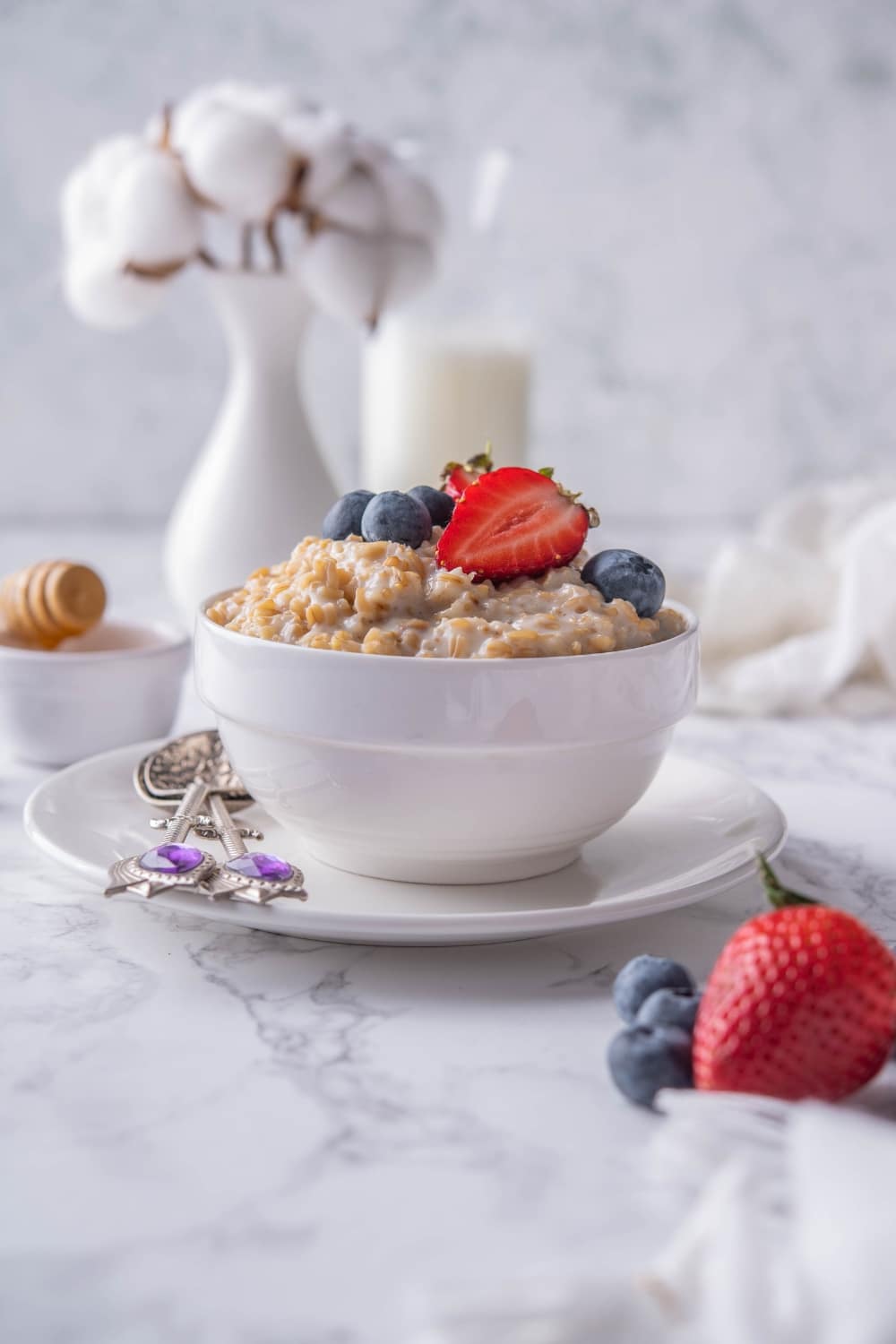 A bowl of steel cut oatmeal garnished with blueberries and sliced strawberries, served over a plate with two dessert spoons.