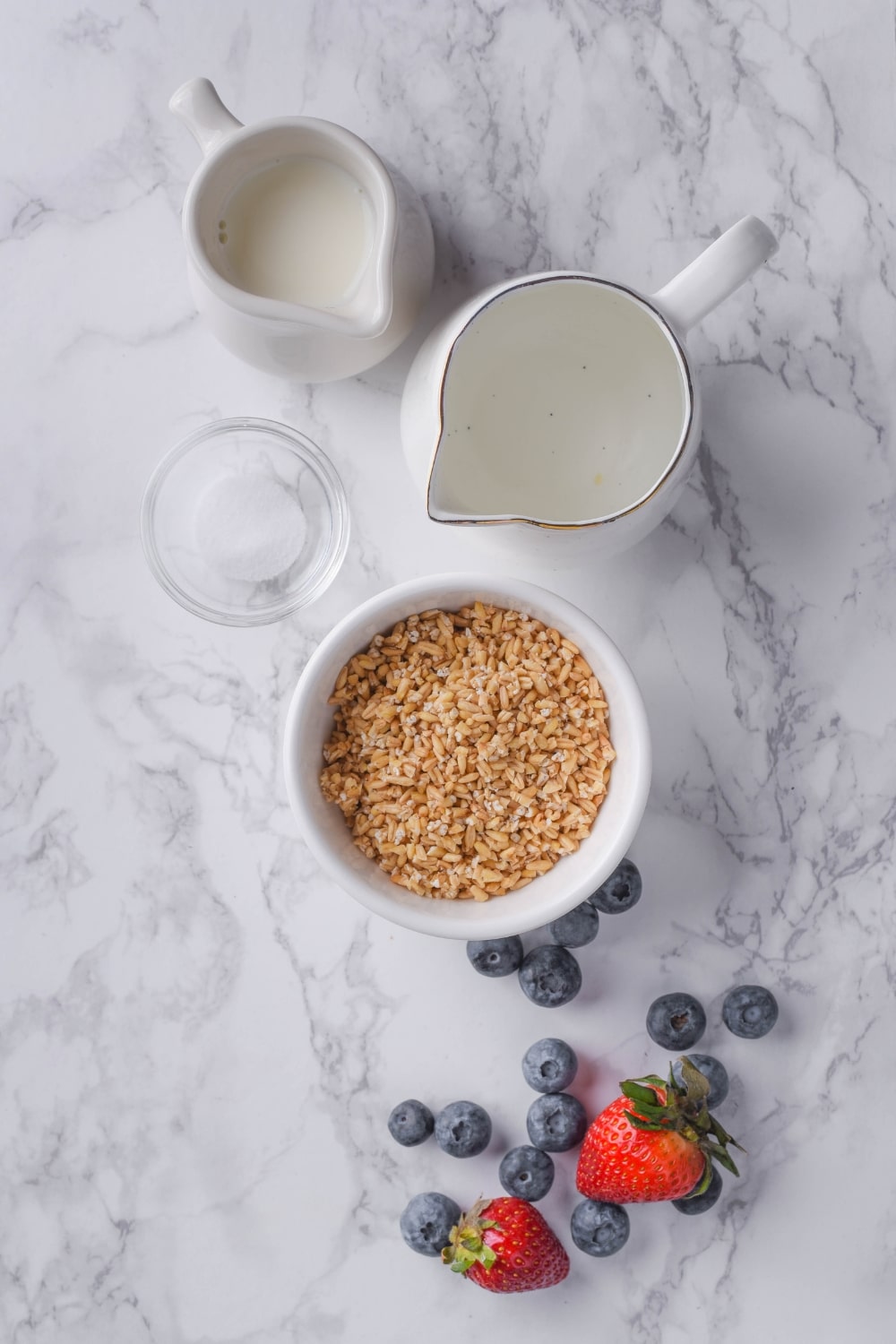 A bowl of uncooked steel cut oats, small ceramic pitchers of water and almond milk, a small glass bowl of salt, and fresh blueberries and strawberries.