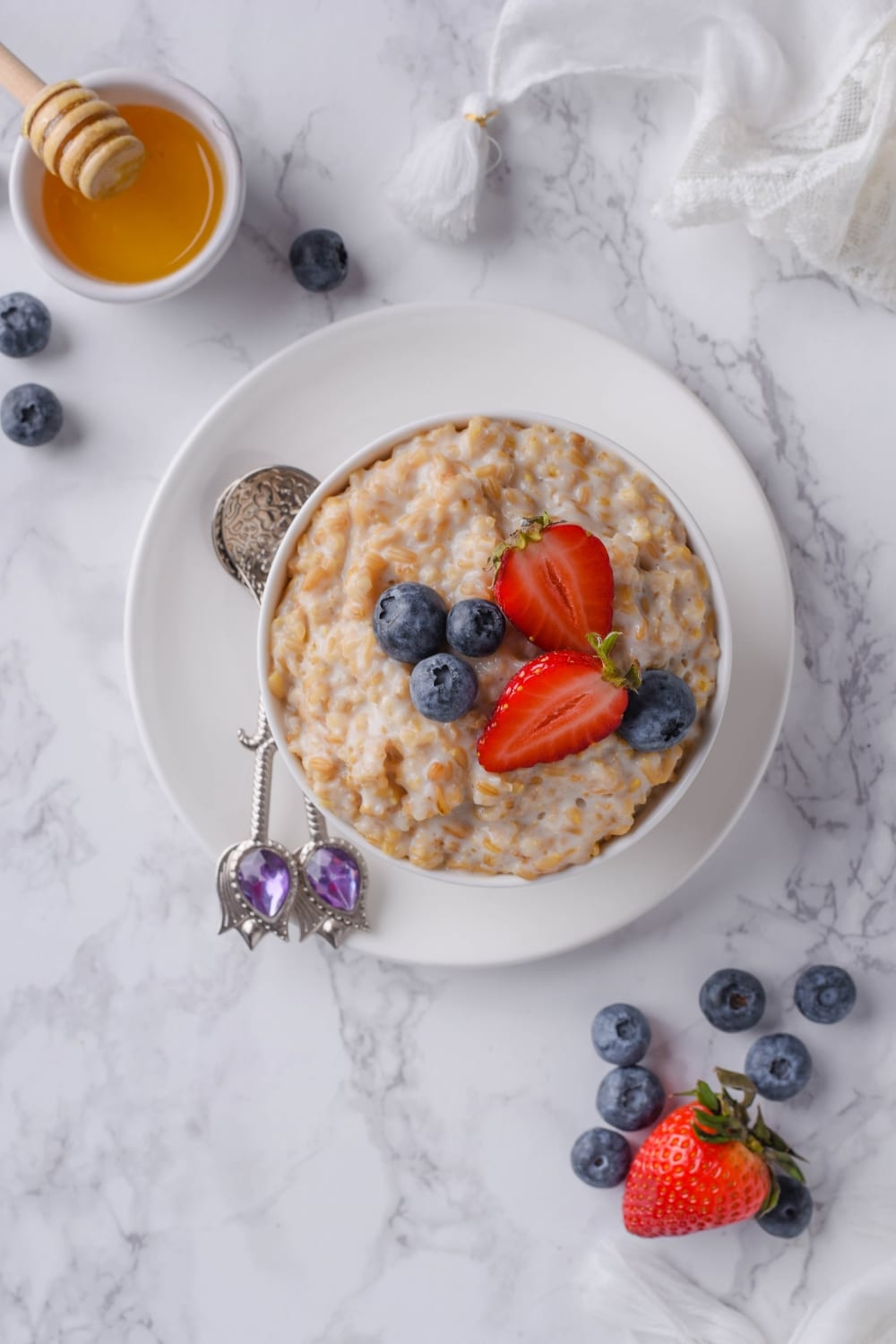 Top view of a bowl of steel cut oats, garnished with blueberries and strawberries, and served with two dessert spoons and a small bowl of honey with a honey dipper.