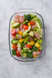 Chopped chicken breasts, broccoli florets, zucchini, bell peppers, and cherry tomatoes covered in olive oil and seasoning in a glass baking dish.