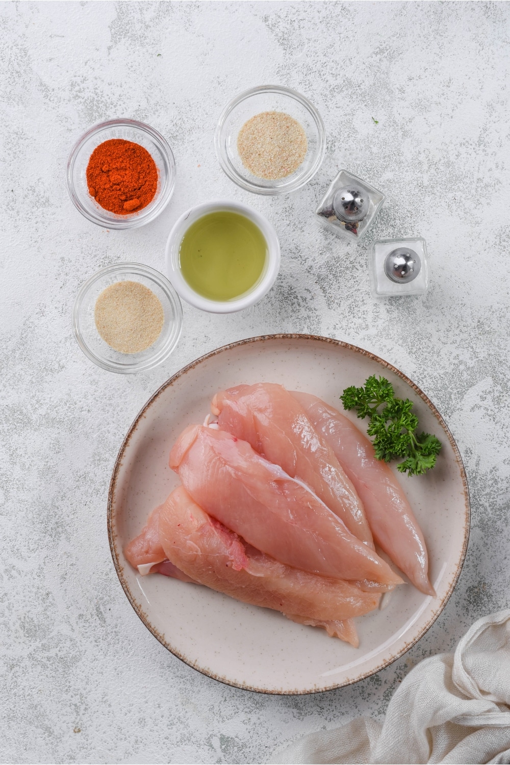 Raw chicken tenders on a plate, small bowls of paprika, garlic powder, onion powder, and olive oil, and salt and pepper shakers.