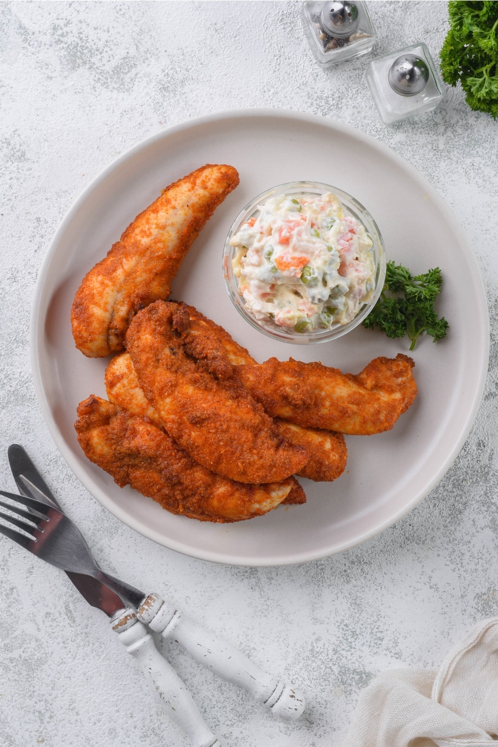 Top view of baked chicken tenders on a plate served with a small bowl of creamy potato salad.