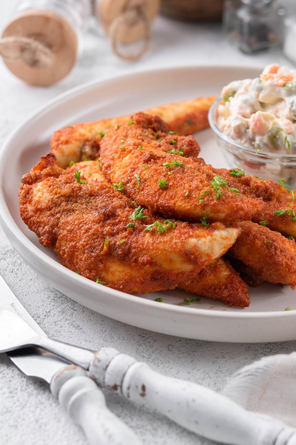 Baked chicken tenders garnished with chopped parsley with a small bowl of creamy potato salad.