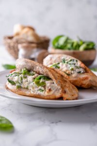Two cream cheese and spinach stuffed chicke breasts on a plate, in front of bowls of bread and spinach.