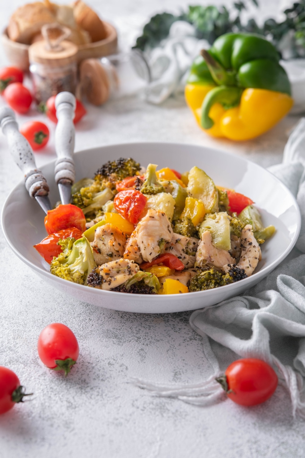 Roasted chicken and veggies served in a shallow white bowl with two forks. Behind are cherry tomatoes, bell peppers, and a bowl of bread.