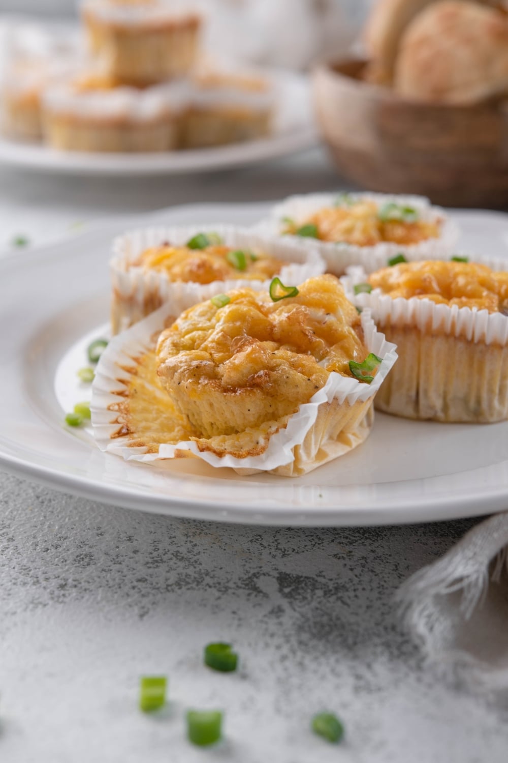 Four sausage egg muffins in muffin liners, garnished with green onions, on a white plate. One muffin has part of its liner peeled open.