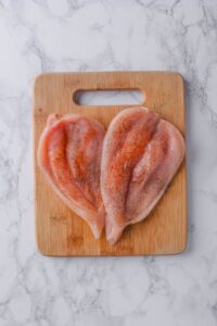 Two chicken breasts sliced open and seasoned with paprika, salt, and pepper on a wooden cutting board.