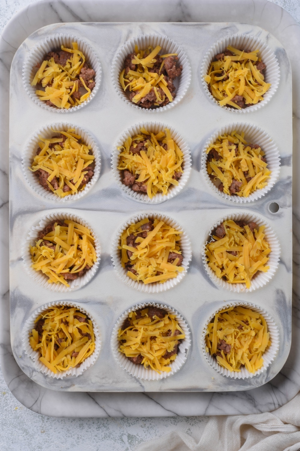 A 12-cup muffin tin with liners filled with crumbled Italian sausage and shredded cheddar cheese.