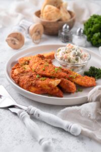 Baked chicken tenders on a plate served with a small bowl of creamy potato salad. Behind is a small bowl of bread.
