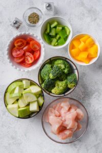 Bowls of chopped raw chicken breast, bell peppers, cherry tomatoes, zucchini, broccoli florets, Italian seasoning, and salt and pepper shakers.