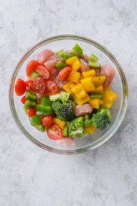 Chopped chicken breasts, broccoli florets, zucchini, bell peppers, and cherry tomatoes covered in olive oil and seasoning in a glass bowl.