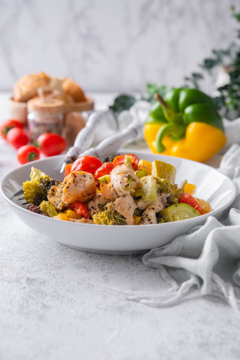 Roasted chicken and veggies on a white plate. Behind are some cherry tomatoes, bell peppers, and a bowl of bread.