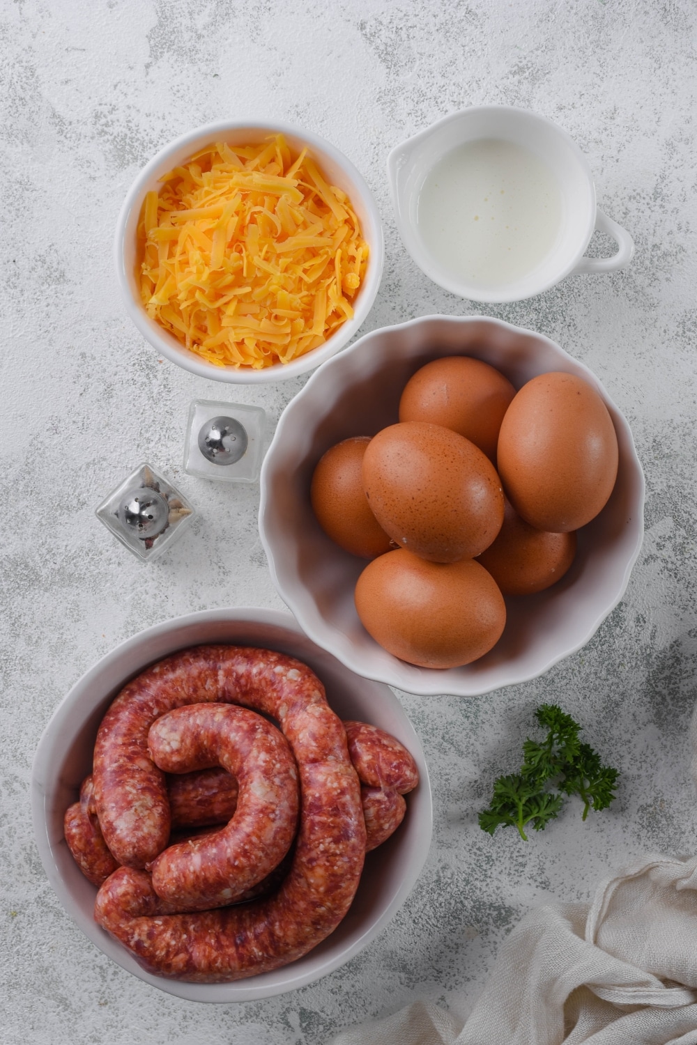 Bowls of Italian sausage, whole eggs, and shredded cheddar cheese, a small pitcher of heavy cream, and salt and pepper shakers.