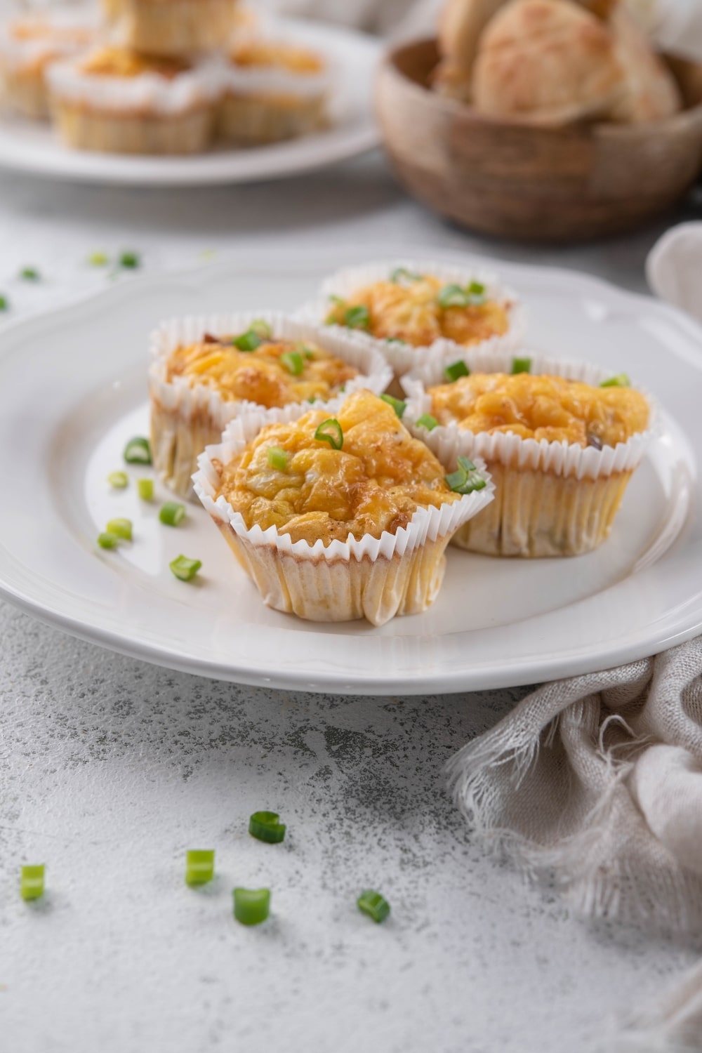 Four sausage egg muffin cups garnished with green onion on a plate. Behind is another plate of stacked sausage egg muffins.