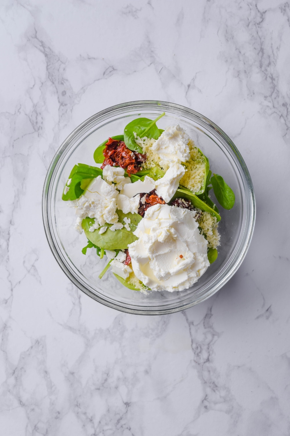 A glass bowl with spinach, cream cheese, feta cheese, chopped sun dried tomatoes, and shredded parmesan.