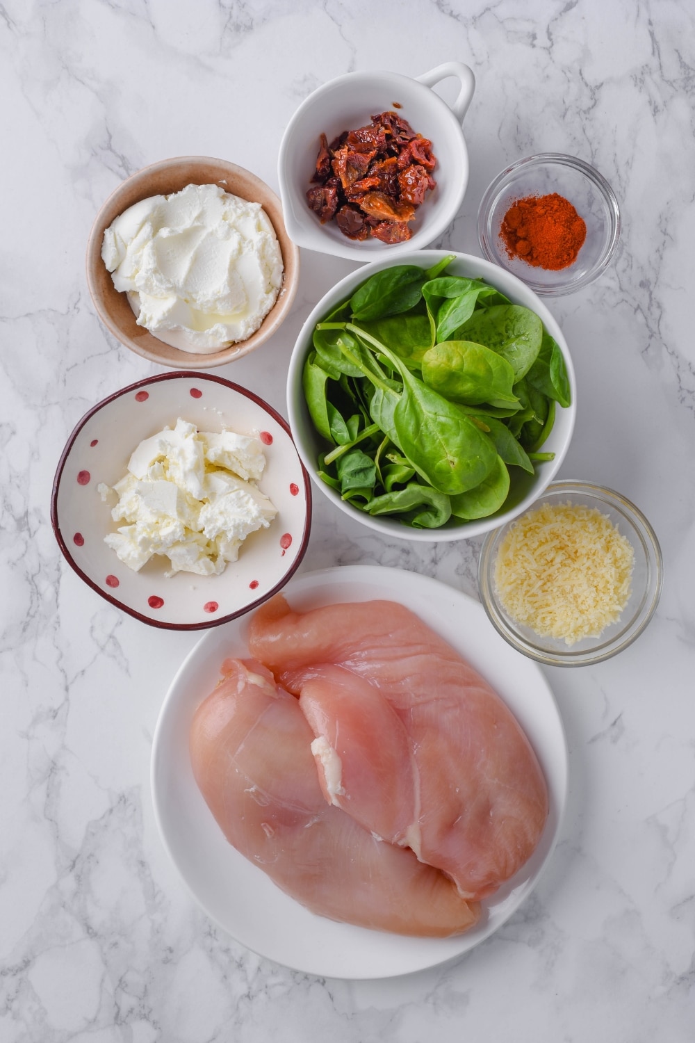 A plate of raw chicken breasts and bowls of spinach, cream cheese, feta cheese, sun dried tomatoes, shredded parmesan, and paprika.