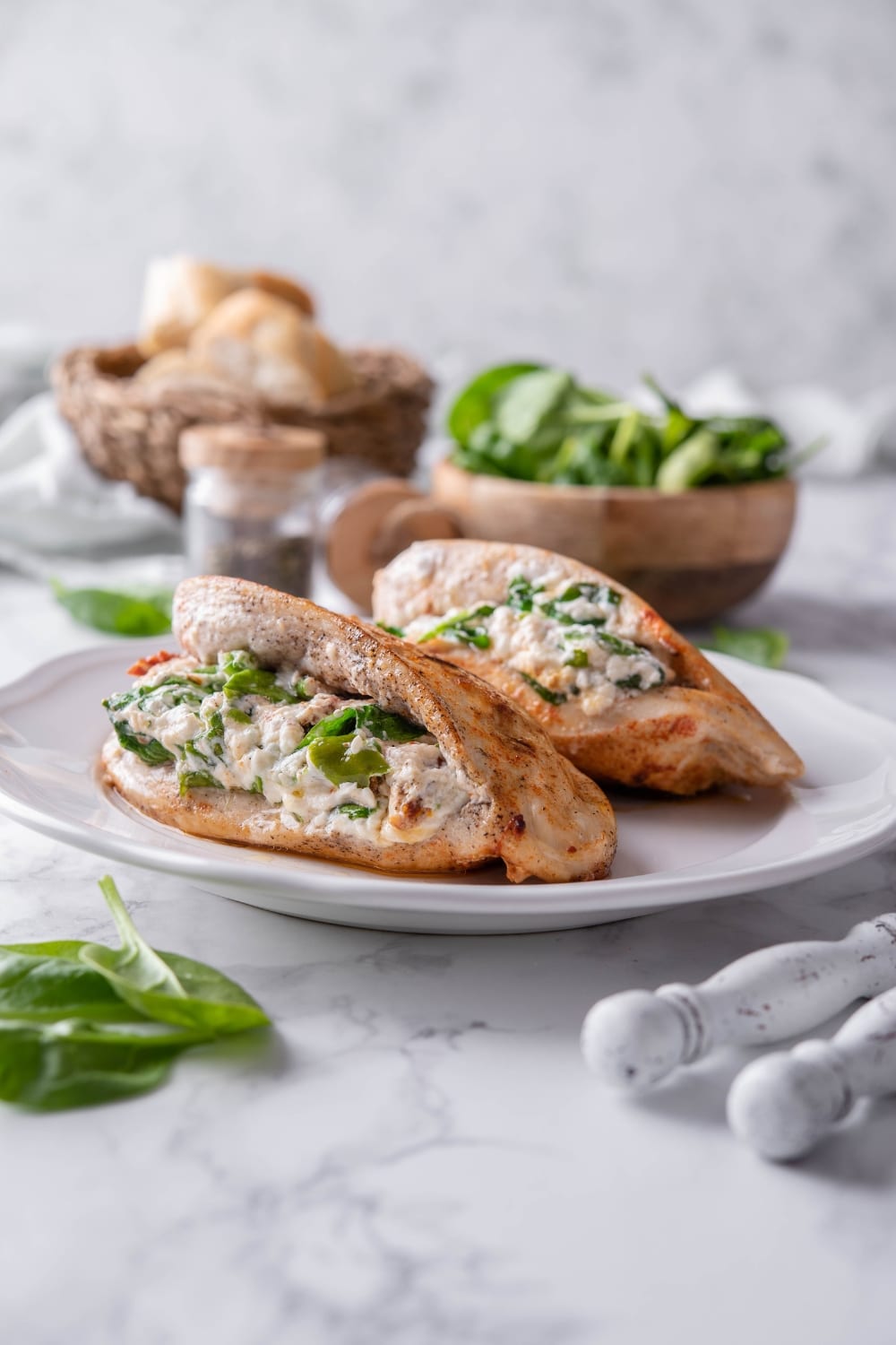 Two stuffed chicke breasts on a plate, served with bowls of bread and spinach.