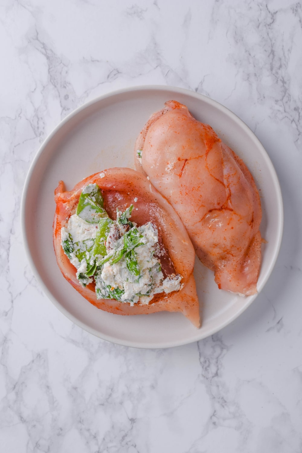 Two uncooked stuffed chicken breasts on a plate, one is open to show the cheese, spinach, and sundried tomato filling.