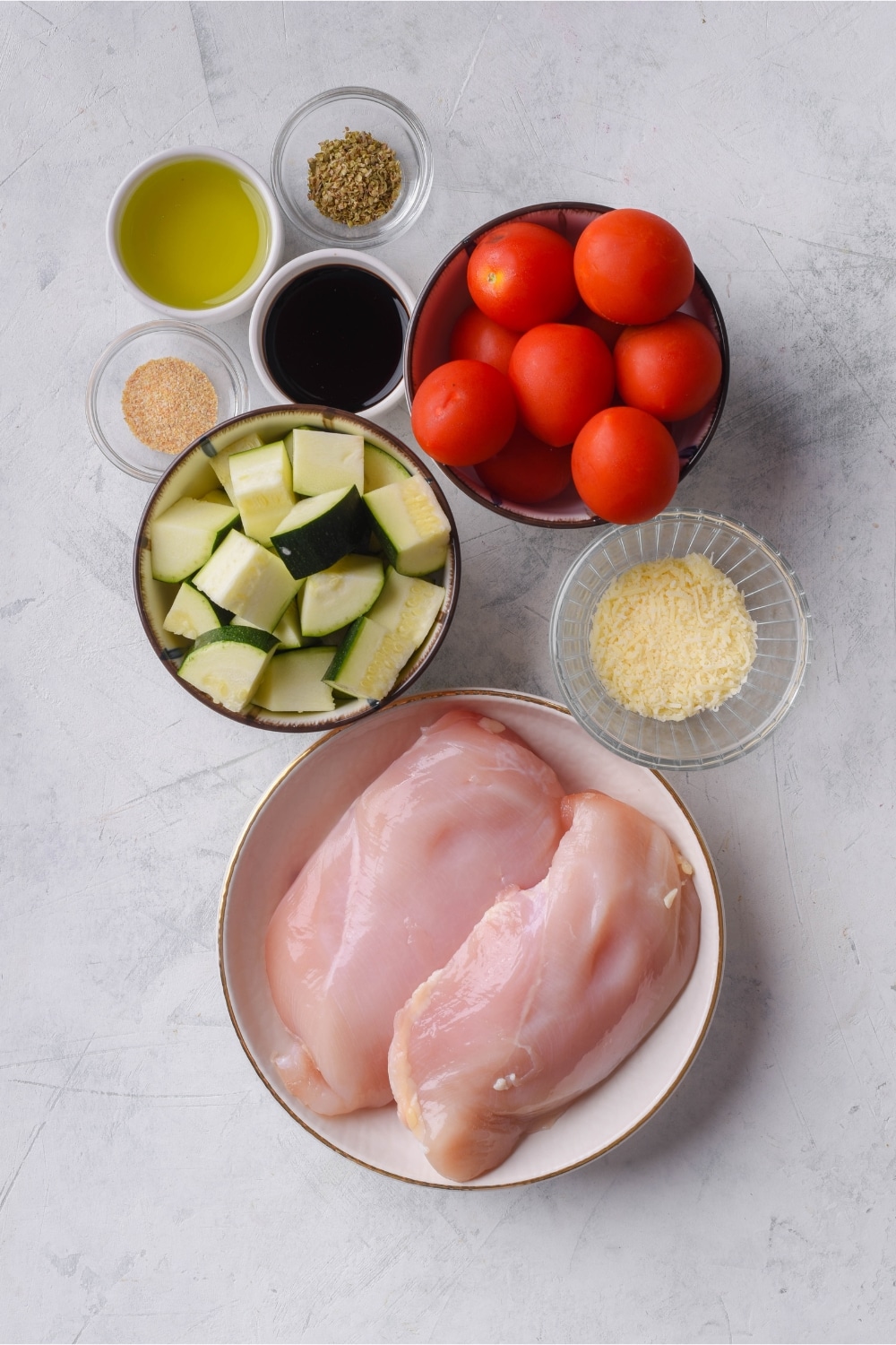 A plate of raw chicken filets, bowls of cherry tomatoes and chopped zucchini, and smaller bowls of seasonings and oil.