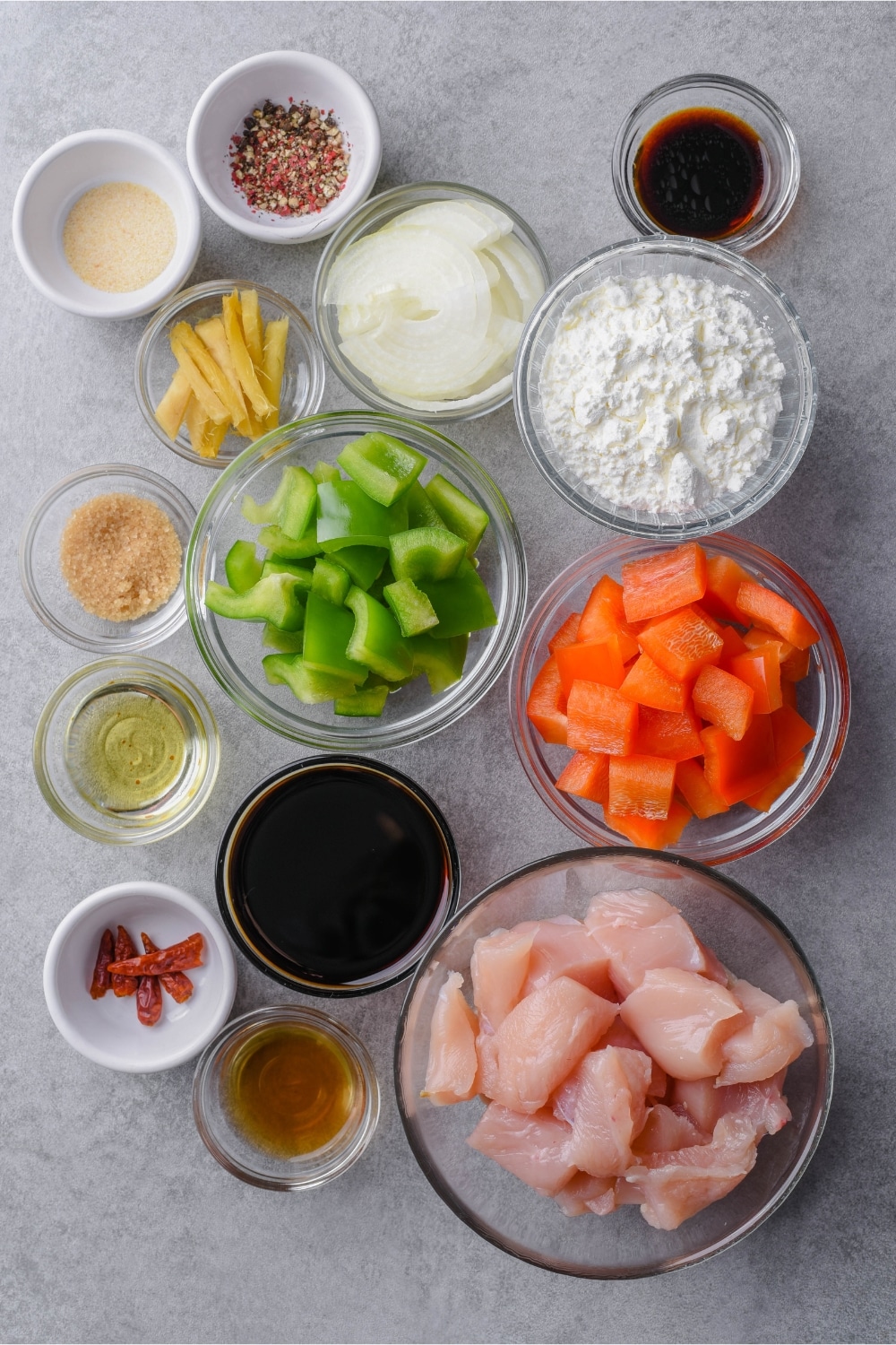 Glass and ceramic bowls of different sizes holding chicke breast chunks, bell peppers, sliced onions, soy sauce, Shaoxing wine, rice vinegar, brown sugar, sesame oil, cornstarch, garlic, dried chilies, ginger, and Sichuan peppercorns.