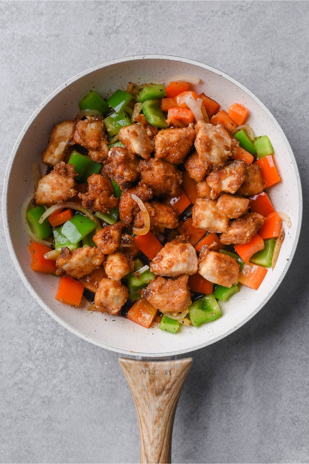 Fried pieces of chicken with sliced bell peppers and onions in a white skillet with a wooden handle.