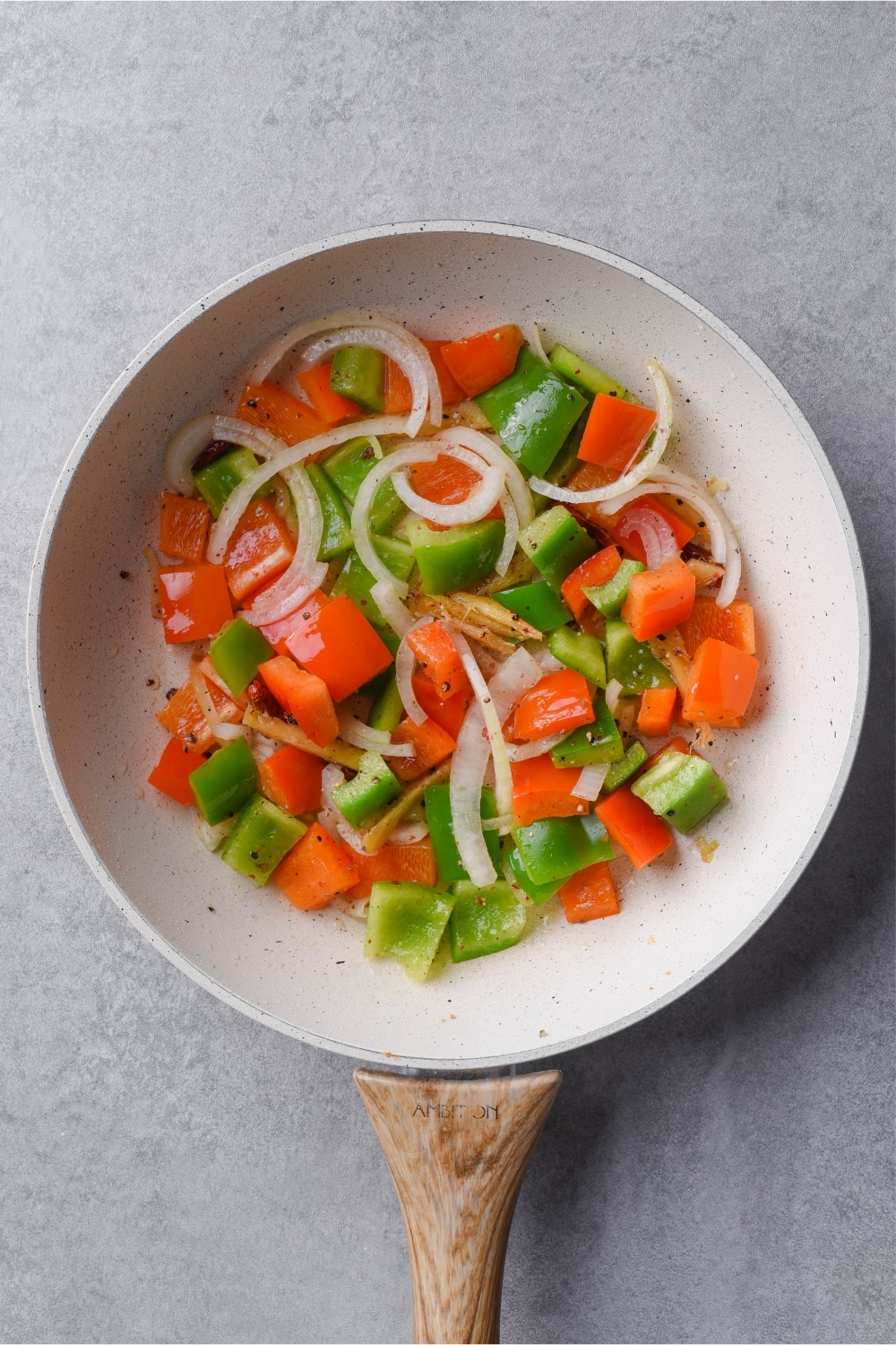 Sliced bell peppers and onions in a white skillet with a wooden handle.