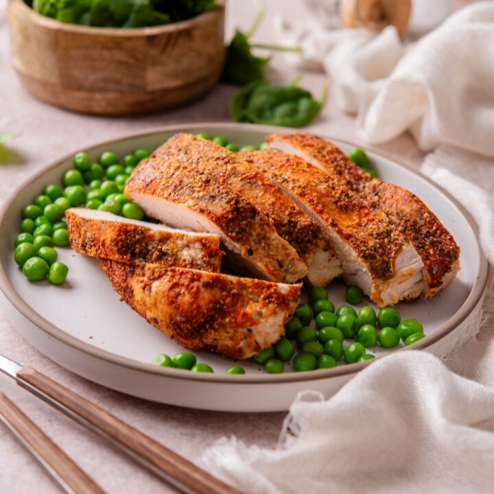 Baked boneless skinless turkey breast on a plate with green peas, next to a bowl of spinach.