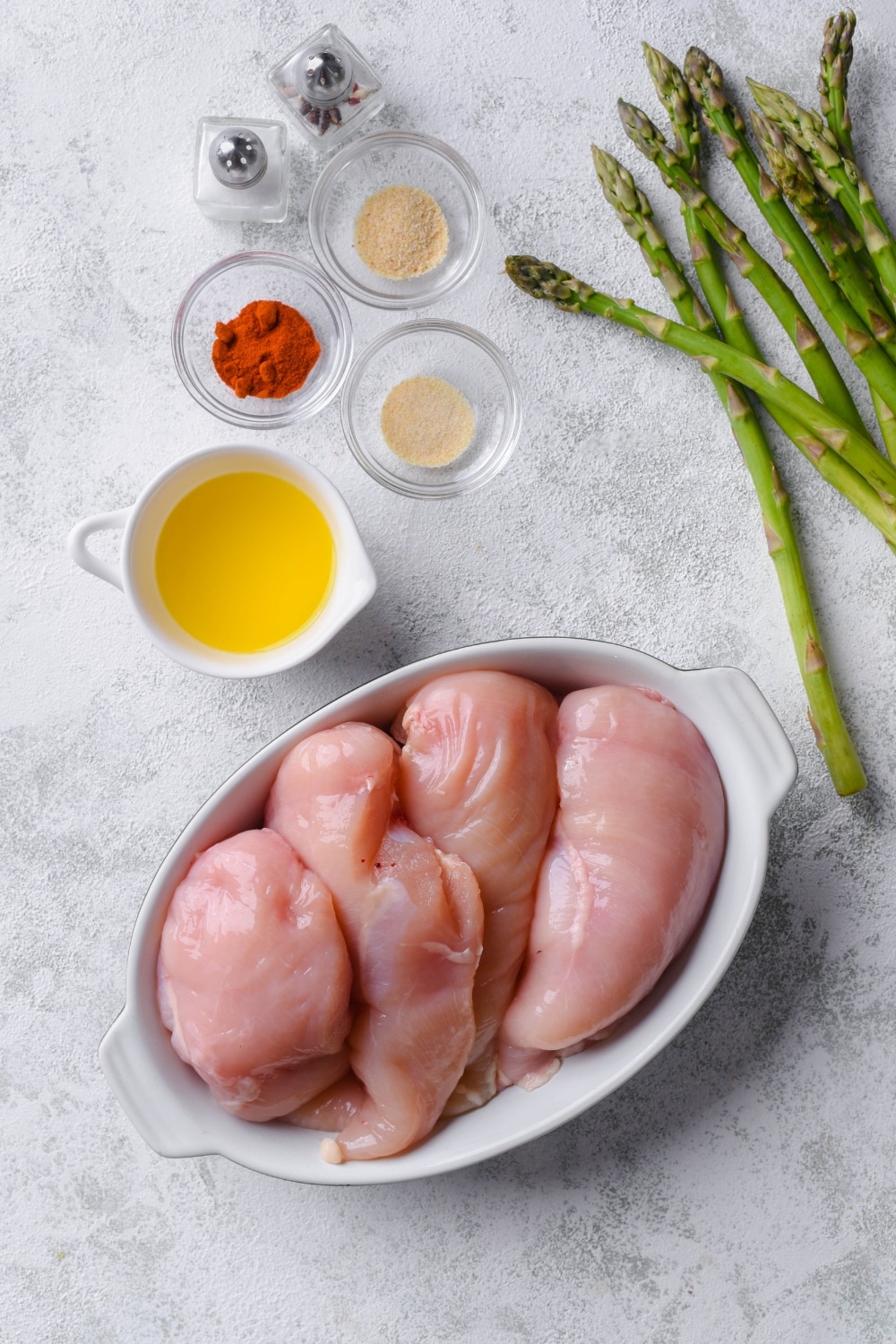 A tray of four raw chicken breasts, a small pitcher of melted butter, glass bowls of seasonings, salt and pepper shakers, and a bunch of asparagus.