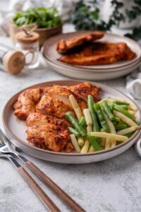 Baked thin chicken breasts and steamed green beans on a plate. Next to it is another plate of baked thin chicken breasts.