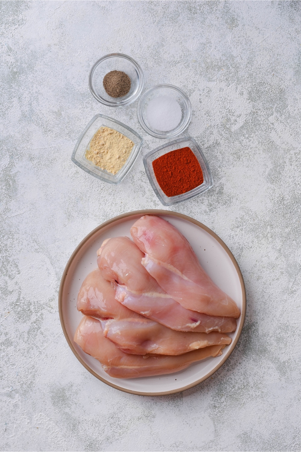 Thin chicken breasts on a plate and small glass containers of garlic powder, paprika, salt, and pepper.