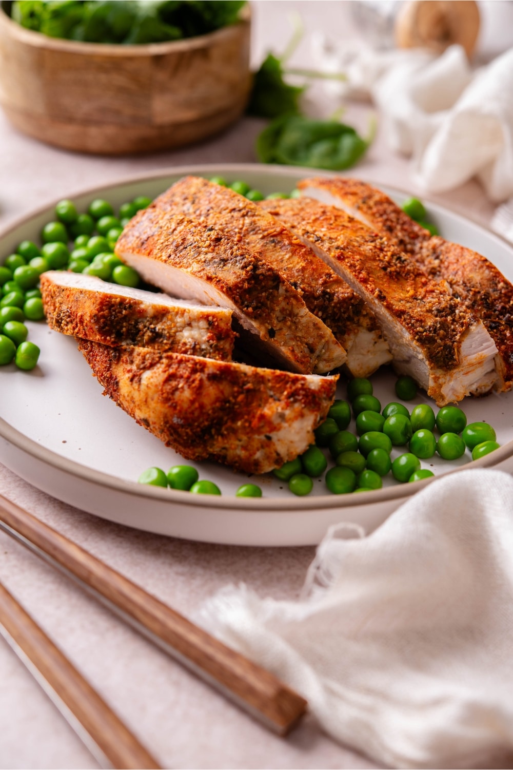 Baked boneless skinless turkey breast with green peas on a plate, next to a bowl of spinach.