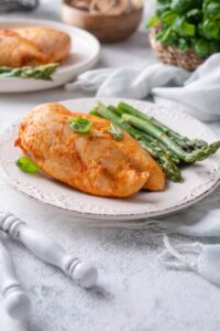A plate of seasoned baked chicken breasts served with asparagus and garnished with basil leaves.