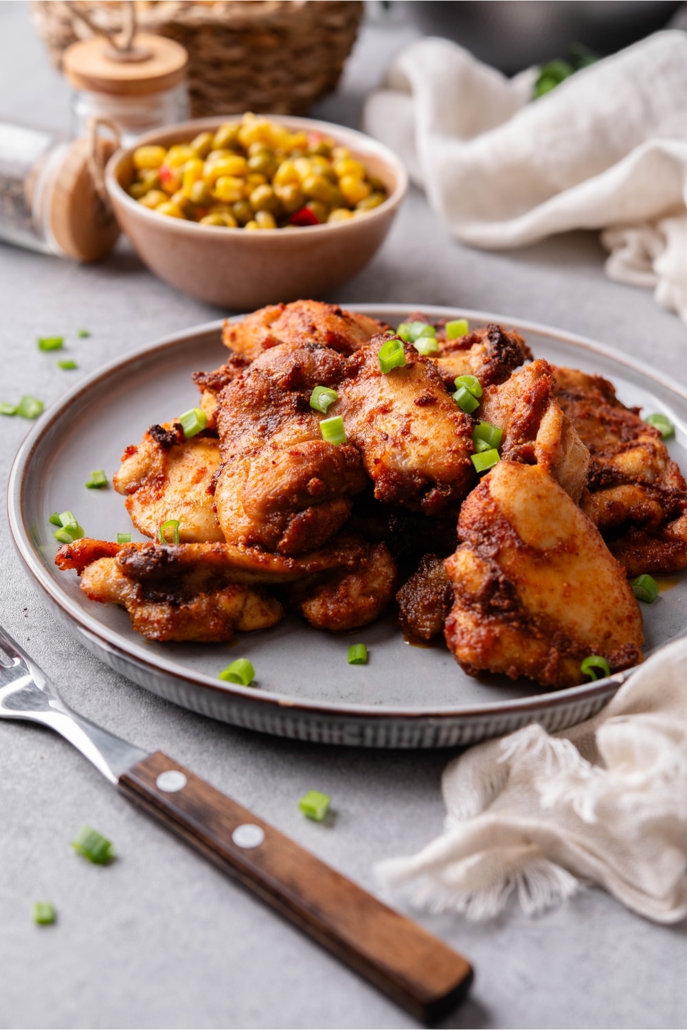 A plate of baked boneless skinless chicken thighs garnished with chopped green onions and served with a small bowl of corn and peas.