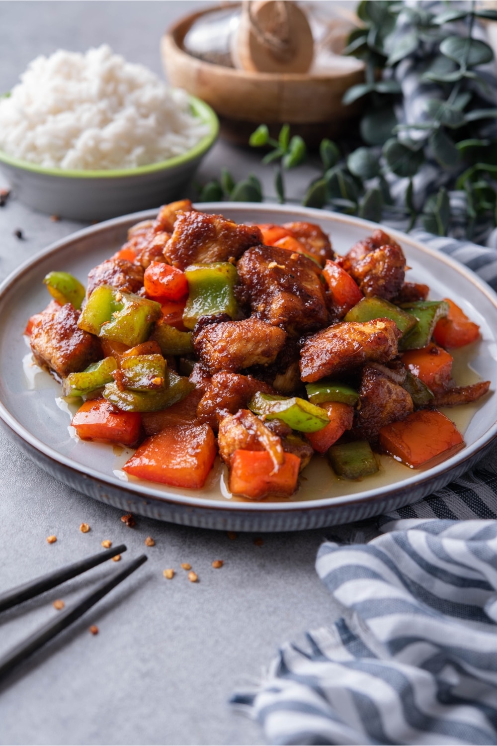 A plate of Szechuan chicken with peppers, served with chopsticks in front of a bowl of rice.