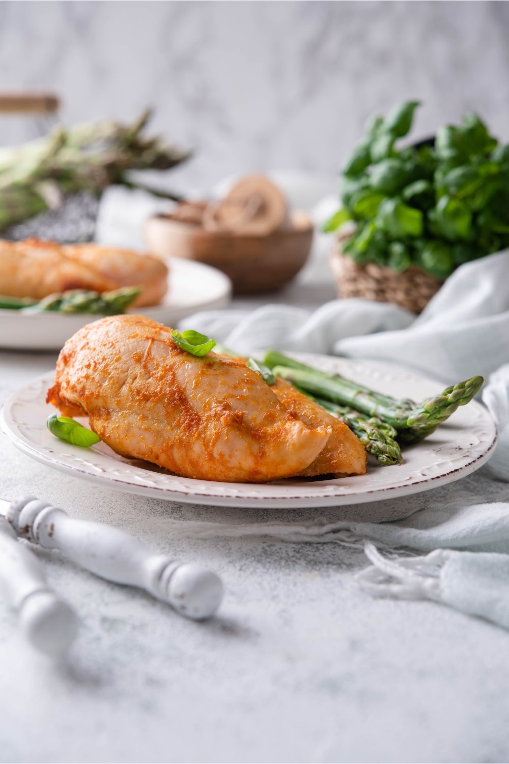 A plate of baked chicken breasts served with asparagus and garnished with basil leaves. A second serving is in the back.