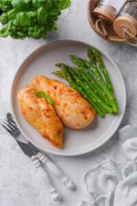 Top view of a plate of baked chicken breasts served with asparagus and garnished with basil leaves.
