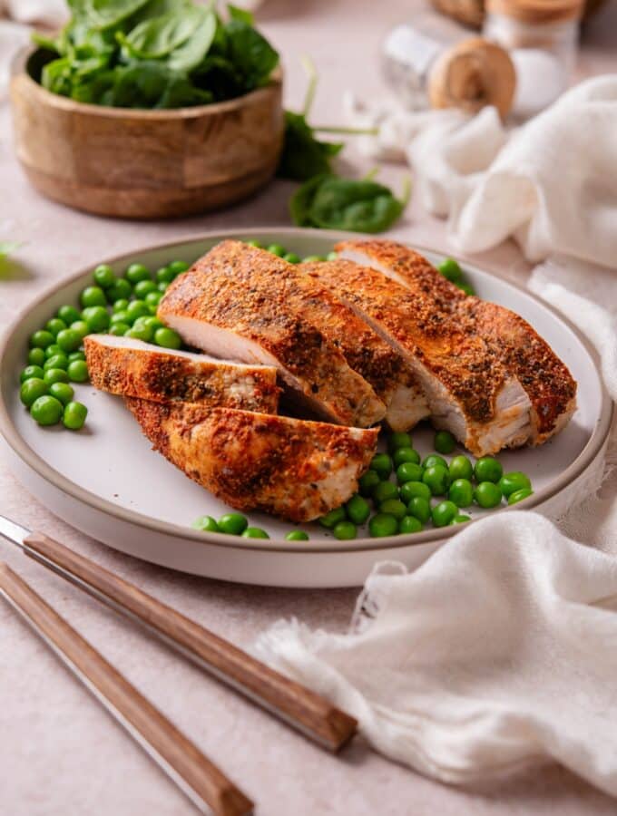 Baked boneless skinless turkey breast and green peas on a plate next to a bowl of spinach.