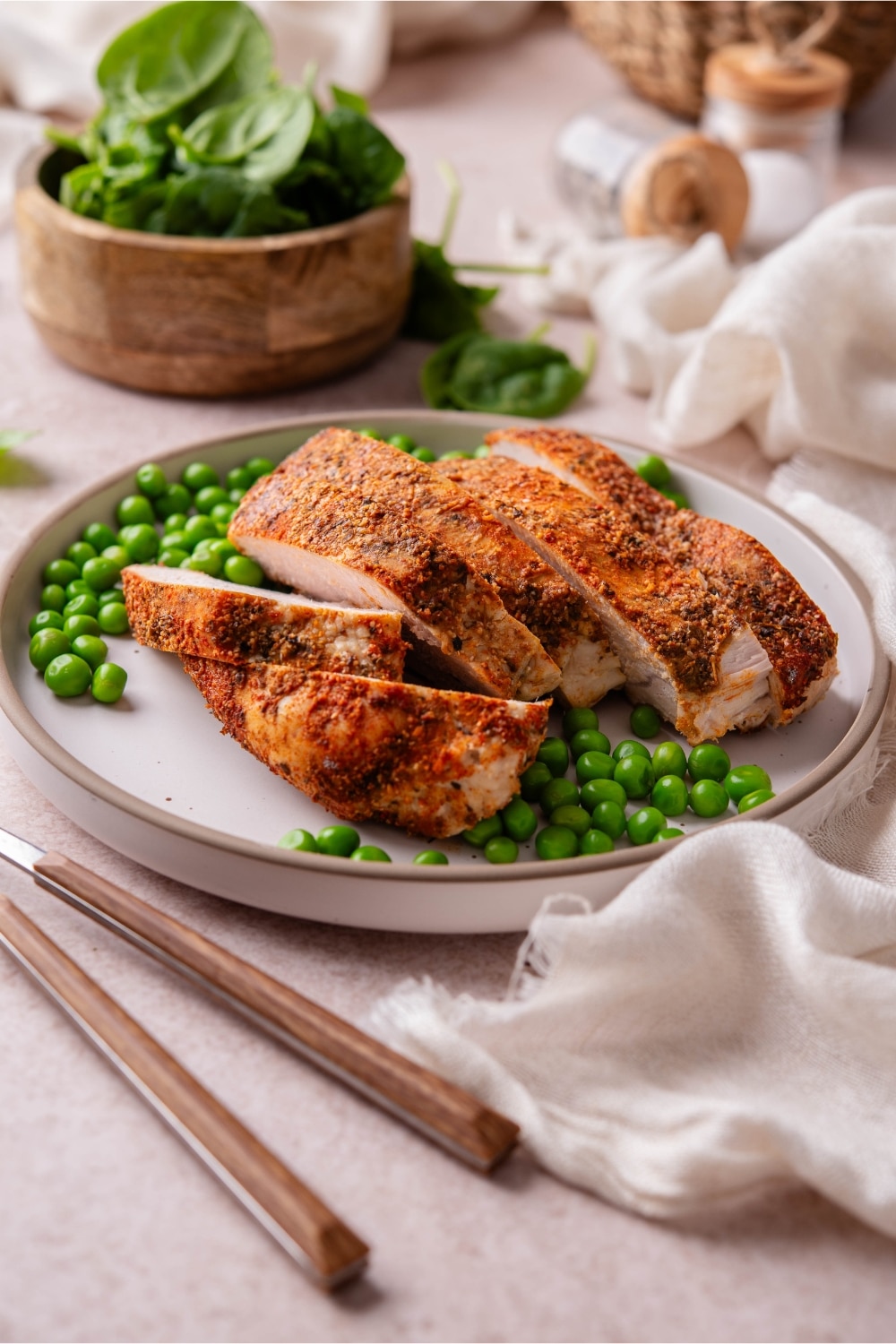 Baked boneless skinless turkey breast and green peas on a plate next to a bowl of spinach.