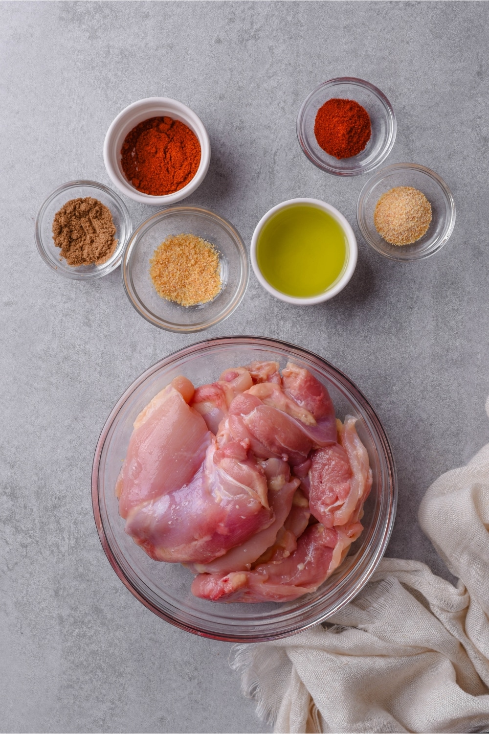 A glass bowl of raw boneless skinless chicken thighs and smaller bowls of spices and oil.