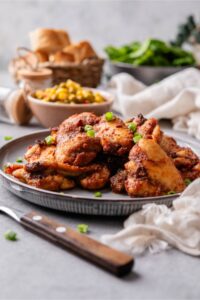 Baked boneless skinless chicken thighs garnished with chopped green onions on a plate and served with a small bowl of corn and peas.