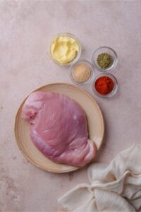 A plate of boneless skinless chicken breast and smaller bowls of spices and butter.