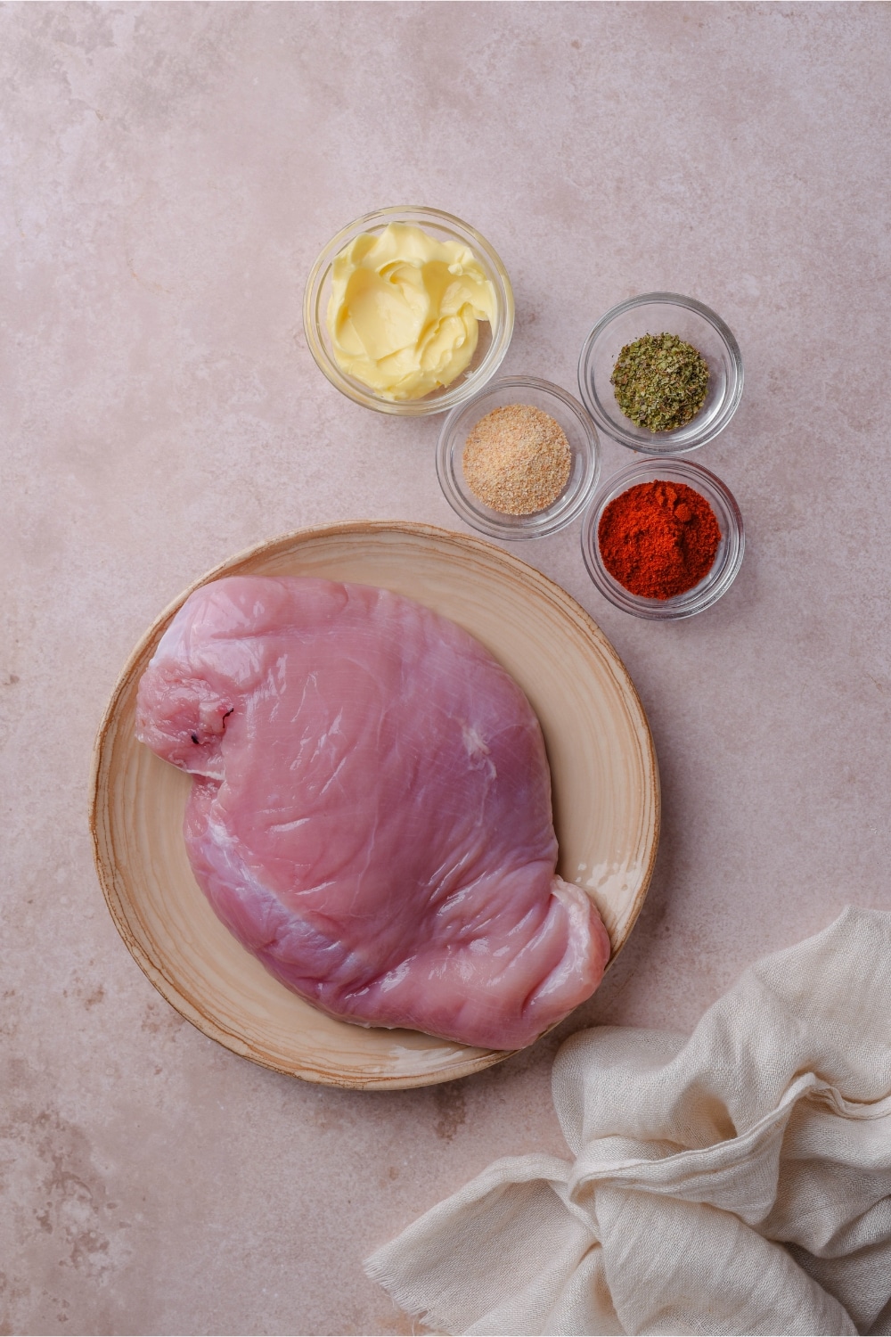 A plate of boneless skinless chicken breast and smaller bowls of spices and butter.