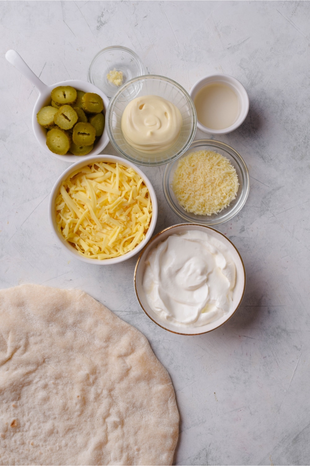 Raw pizza dough and small bowls of dill pickles, mozzarella, parmesan, minced garlic, sour cream, mayo, and lemon juice.