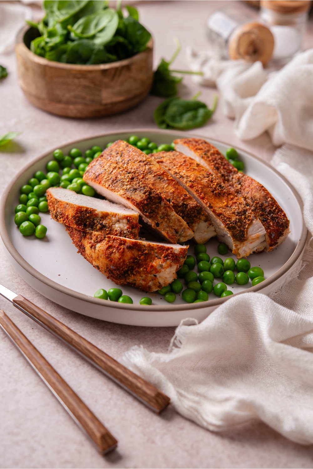 Sliced baked turkey breast on a plate with green peas next to a bowl of spinach.