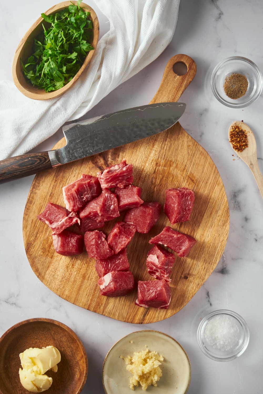 Steak bite cubes on a round cutting board with a knife, surrounded by bowls of parsley, butter, minced garlic, and pepper, and a spoon of red pepper flakes.